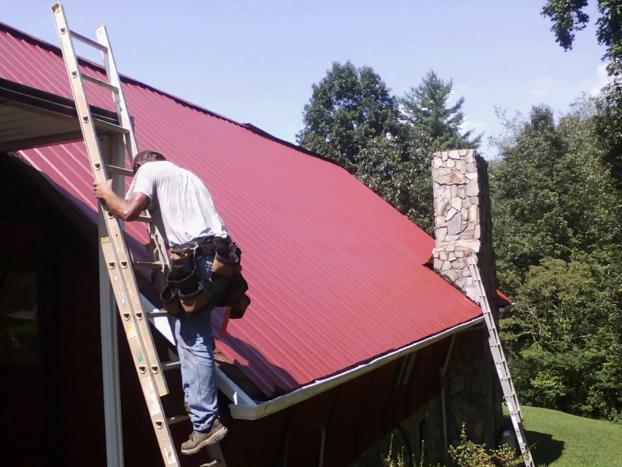 Roofer on a ladder beside a red metal roof and stone chimney — Soffit & Fascia Repair by Roofing Brothers in Lafayette